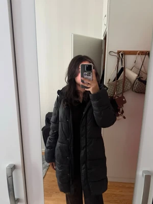 Svart lång dunjacka med huva - Säljer en svart lång dunjacka med huva och dragkedja framtill. Jackan har en quiltad design och är perfekt för kalla vinterdagar. Passar dig som vill ha en stilren och varm jacka med enkel look.