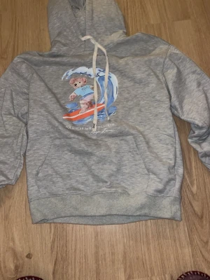 Grå hoodie Polo Ralph Lauren björn - Säljer en grå hoodie från Polo Ralph Lauren med en surfande björn och vågor på bröstet. Hoodien har huva med vita snören och en stor magficka. Mjuk och skön bomullsblandning, perfekt för chill dagar. Klassisk streetwear-vibe med unik print.