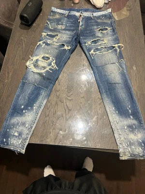 Blå slitna jeans med stora hål - Säljer ett par blå jeans med kraftiga slitningar och stora hål på både fram- och baksida. Jeansen har en tvättad look med ljusa partier och spridda färgstänk. Modellen är rak och har klassiska fem fickor. Perfekt för dig som gillar en edgy och trendig stil.