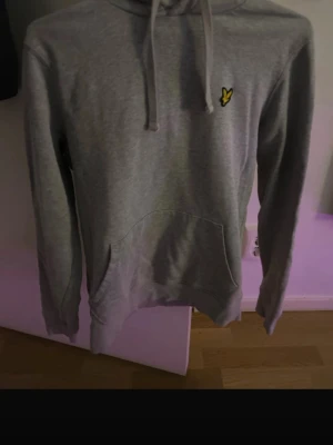 Grå hoodie - Säljer en stilren grå hoodie med huva och dragsnören. På bröstet sitter en liten gul logga. Hoodien har en stor magficka och ribbade muddar vid ärmar och nederkant. Perfekt för en avslappnad och trendig look.