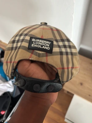 Burberry beige rutig keps - Säljer en klassisk beige keps från Burberry med det ikoniska rutmönstret i svart, vitt och rött. Kepsen har justerbart spänne bak i svart läder och en böjd skärm. Perfekt för dig som vill ha en stilren accessoar med lyxig känsla.