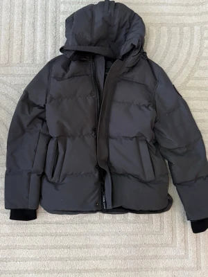 Canada gosse jacka  - Snygg svart dunjacka med stor huva och dragkedja framtill. Jackan har två stora fickor på sidorna och bred kanaler för extra värme. Perfekt för kalla vinterdagar och har en modern, puffig look.