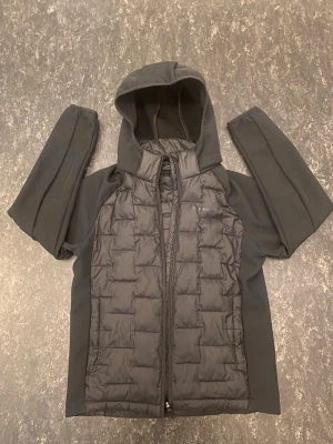 Svart vindjacka med huva - Snygg svart vindjacka med vadderad framsida och mjuka ärmar. Jackan har dragkedja framtill och en praktisk huva. Perfekt för blåsiga dagar och enkel att matcha med olika outfits. Materialet är en mix av syntet och vaddering för extra komfort.
