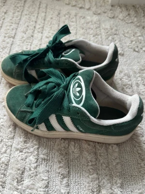 Gröna Adidas Campus sneakers - Säljer ett par gröna Adidas Campus sneakers i mocka med klassiska vita ränder på sidorna och Adidas-logga på plösen. Skorna har breda gröna skosnören och vit sula med gummiyta undertill. Perfekta för dig som gillar retrostil och streetwear.