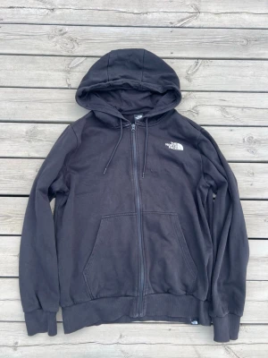 Svart hoodie från The North Face - Säljer en svart hoodie från The North Face med dragkedja, huva med dragsko och två stora fickor framtill. Klassisk logga i vitt på bröstet. Perfekt för en chill och avslappnad stil.