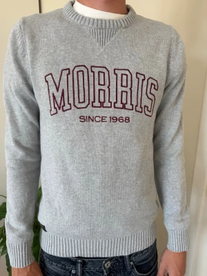 Grå Morris tröja med brodyr - Snygg ljusgrå stickad tröja från Morris med mörkröd broderad logga och texten 'Since 1968' på bröstet. Tröjan har rund halsringning, ribbade muddar och är perfekt för en avslappnad men stilren look. Aldrig använd.