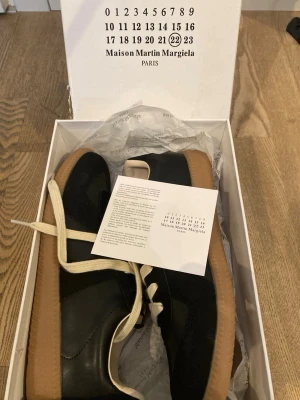 Maison Margiela svarta sneakers - Svarta sneakers från Maison Martin Margiela med ovandel i både mocka och läder, vita snören och klassisk gummisula i brunt. Snygg retrostil med låg profil och diskreta detaljer. Perfekta för dig som gillar minimalistisk och stilren design.