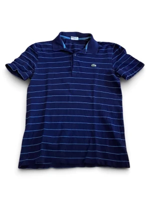 Marinblå randig piké från Lacoste - Snygg marinblå pikétröja från Lacoste med tunna ljusblå ränder och klassisk krage. Tröjan har korta ärmar och den ikoniska krokodilloggan på bröstet. Perfekt för en avslappnad men stilren look.