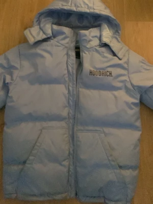 Ljusblå pufferjacka från Hoodrich - Ljusblå pufferjacka från Hoodrich med stor logga på ryggen och mindre logga på bröstet. Jackan har huva, dragkedja framtill och två stora fickor. Perfekt för kalla vinterdagar tack vare det tjocka materialet och den vadderade designen. Men zip är sönder men det går att drar up