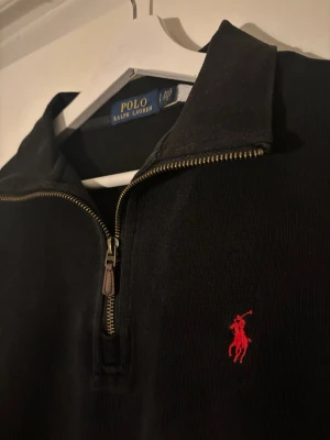 Ralph Lauren Quarter Zip - Svart långärmad tröja från Polo Ralph Lauren med klassisk quarter zip och hög krage. Tröjan har den ikoniska röda broderade hästen på bröstet och är tillverkad i mjukt material. Perfekt för en clean och stilren look.