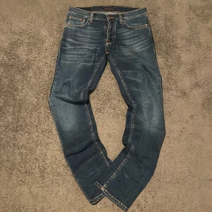 Blå Nudie Grim Tim - Säljer ett par snygga blå jeans från Nudie Jeans Co med snygga slitningar och orangea sömmar. Jeansen har fem fickor, normal passform och är tillverkade av ekologisk bomull. Bra potential för snygga fades.