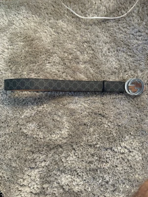 Märkes bälte - Snyggt svart bälte med diskret mönster och stor rund silverfärgad spänne med GG-detalj. Bältet har en klassisk design och insidan är i ljusbrunt material. Perfekt för att lyfta vilken outfit som helst.