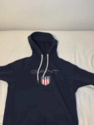 Mörkblå hoodie från GANT med logga - Snygg mörkblå hoodie från GANT med vit dragsko i huvan och stor GANT-logga samt sköld med stjärnor och ränder på bröstet. Klassisk passform och mjukt material, perfekt för en avslappnad stil.