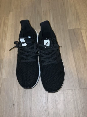 Svarta Adidas sneakers med snörning - Säljer ett par svarta Adidas Ultraboozt sneakers med klassisk vit sula och svart ovandel i mesh. Skorna har snörning och diskret Adidas-logga på plösen. Perfekta för dig som gillar stilrena och sportiga sneakers.