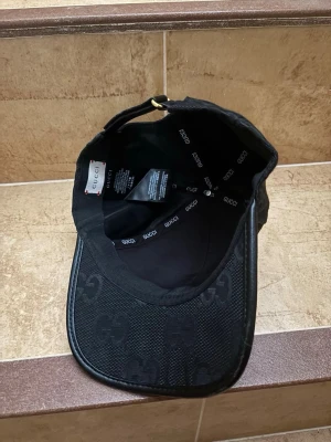 Svart keps från Gucci med GG-mönster - Snygg svart keps från Gucci med det klassiska GG-mönstret över hela kepsen. Justerbar rem baktill med metallspänne och böjd skärm. Tillverkad i ett mesh-liknande material som ger en exklusiv känsla och stilren look.
