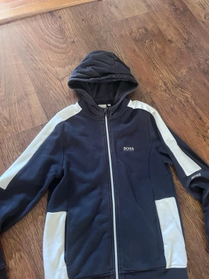 Svart hoodie med vita detaljer från BOSS - Snygg svart hoodie från BOSS med vita paneler längs ärmar och sidor. Dragkedja framtill, huva och diskret BOSS-logga på bröstet och ärmen. Perfekt för en avslappnad och sportig stil.