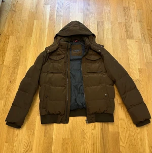 Pufferjacka - Säljer en brun pufferjacka med avtagbar huva och dragkedja framtill från Tommy Hilfiger. Jackan har två bröstfickor med lock och två sidofickor med knappstängning. Den har använts några få gånger!