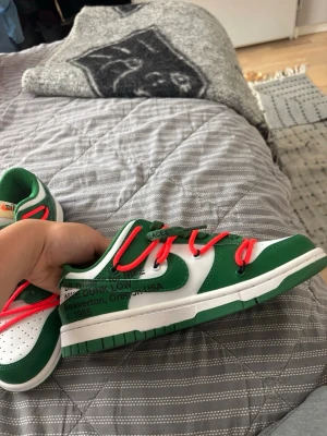 Nike Dunk Low Off-White Grön/Vit - Nike Dunk Low Off-White sneakers i grönt och vitt läder med dubbla snörningar – gröna och orangea. Klassisk låg modell med perforerad tåbox och Off-White-typisk text på sidan. Snygg och unik design som sticker ut.