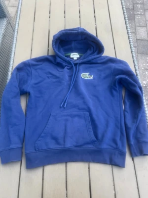 Blå hoodie från Lacoste med huva - Snygg blå hoodie från Lacoste med klassisk krokodillogga på bröstet. Hoodien har dragsko i huvan och en stor magficka framtill. Perfekt för en avslappnad och sportig stil. Materialet känns mjukt och bekvämt mot huden.
