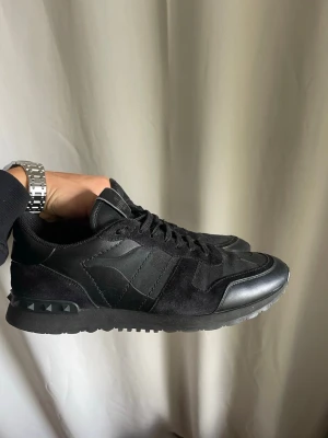Valentino Rockrunner  - Säljer ett par svarta sneakers från Valentino med coola detaljer i mocka och skinn. Skorna har snörning, diskret kamouflagemönster och nitar på hälen. Perfekta för dig som vill ha en stilren men edgy look.