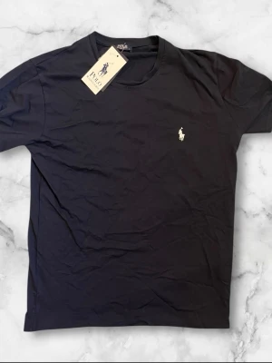 Svart t-shirt från Polo Ralph Lauren - Klassisk svart t-shirt från Polo Ralph Lauren med rund hals och korta ärmar. T-shirten har den ikoniska broderade loggan i vitt på bröstet och är tillverkad i mjuk bomull för en skön känsla.