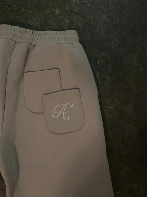 Beige mjukisbyxor med broderad ficka - Beiga mjukisbyxor med raka ben, resår i midjan och dragsko. Två sidofickor och två dekorativa fickor bak, varav en har ett broderat A och en stjärna. Mjukt material, perfekt för chill dagar.