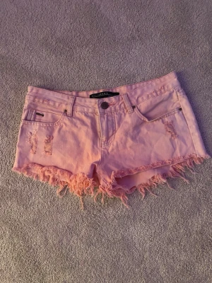 Ljusrosa fransiga jeansshorts - Säljer ett par ljusrosa lågmidjade jeansshorts i en stil som trendar mycket och är väldigt eftertracktad, de har fransig kant och slitna detaljer💕Perfekta för sommaren, säljer pga att de inte kommit till användning❤️