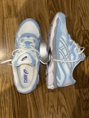Asics sneakers i blått och vitt - Snygga Asics sneakers i ljusblått och vitt med klassisk logga på sidan. Skorna har snörning och är tillverkade i en kombination av mesh och syntetmaterial för en sportig look. Perfekta för dig som gillar stilrena och bekväma sneakers.