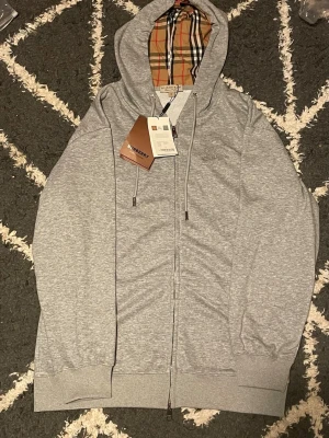 Grå zip hoodie från Burberry - Snygg grå zip hoodie från Burberry med klassiskt rutigt foder i huvan och dragsko. Helt ny, pris kan diskuteras därför tar jag gärna emot prisförslag.OBS EXTRA OVERSIZED!!!