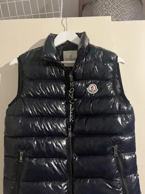 Svart dunväst från Moncler - Säljer en snygg svart Moncler-väst i klassisk blank puffer-modell. Stilren och lätt att matcha.  Skick: Bra skick, sparsamt använd Färg: Svart (glansig)  Detaljer:  * Dragkedja framtill * Två sidofickor med zip * Moncler-logga på bröstet * Hög krage  OBS: Jag vet inte om den är äkta, men den går att skanna och leder till Monclers hemsida.                      Där av priset😁