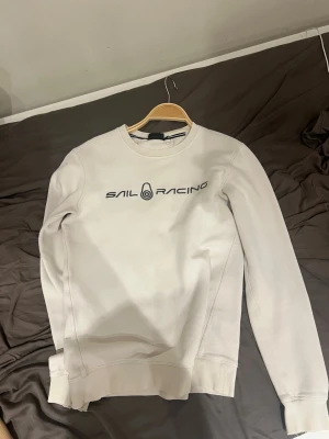 Vit sweatshirt från Sail Racing - Säljer en stilren vit sweatshirt från Sail Racing med svart logotyptryck på bröstet. Tröjan har rund halsringning, långa ärmar och ribbade muddar vid ärmslut och nederkant. Perfekt för en clean och sportig look.