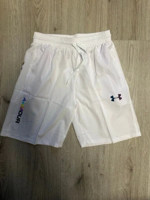 Vita shorts från Under Armour - Snygga vita shorts från Under Armour med elastisk midja och snörning. Shortsen har logga i regnbågsfärger på ena benet och klassisk UA-logga på andra. Perfekta för träning eller chill dagar. Tillverkade i ett lätt och bekvämt syntetmaterial.