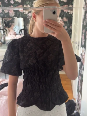 Svart blommig blus med puffärm - Snygg svart blus med blommigt mönster och puffiga korta ärmar. Toppen har smockad midja som ger en markerad siluett och rund halsringning. Perfekt för dig som vill ha en trendig och feminin look.