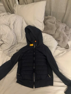 parajumper  - Snygg mörkblå pufferjacka från Parajumpers med vadderad front, mjuka ärmar och huva. Jackan har dragkedja framtill, två fickor med dragkedja och detaljer i orange. Perfekt för kalla vinterdagar och riktigt skön att bära.