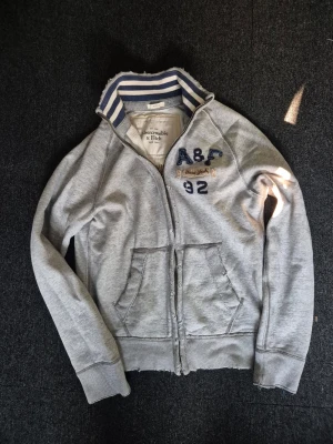Grå zip-hoodie Abercrombie & Fitch - Säljer en sjukt snygg zip-up tröja från Abercrombie & Fitch. Storleken är M och passar ca 175-185. Tröjan har en sliten design från fabrik som ger den en unik vintage look. Tröjan passar perfekt nu till hösten! Denna modell tillverkas inte längre och är därför mycket sällsynt! Priset kan diskuteras vid snabb affär!😊
