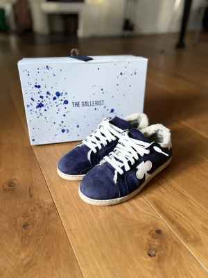 The gallerist winter edition - Mörkblå sneakers i mocka med vit sula och vita skosnören. Insidan är fodrad med mjuk vit fuskpäls och skon har en vit detalj på sidan samt vid hälen. Perfekt för dig som gillar unika och bekväma sneakers.