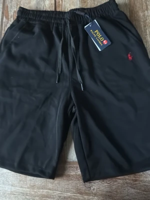 Svarta shorts från Polo Ralph Lauren - Svarta shorts från Polo Ralph Lauren med röd broderad logga på benet. Shortsen har elastisk midja med snörning för skön passform och är tillverkade i ett mjukt material som passar perfekt till sommaren.