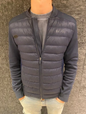 Mörkblå quiltad cardigan från BOSS - Snygg mörkblå cardiganjacka från BOSS med quiltad front och släta ärmar. Jackan har dragkedja framtill, diskret logga på bröstet och ribbade muddar. Perfekt för lager på lager och ger en sportig men stilren look.