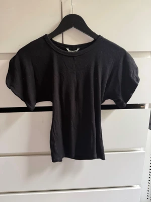 Svart ribbad t-shirt med puffärm - Snygg svart t-shirt med ribbad struktur och puffiga korta ärmar. Perfekt till jeans eller kjol för en trendig look. Materialet känns mjukt och stretchigt, vilket gör den bekväm att bära hela dagen.