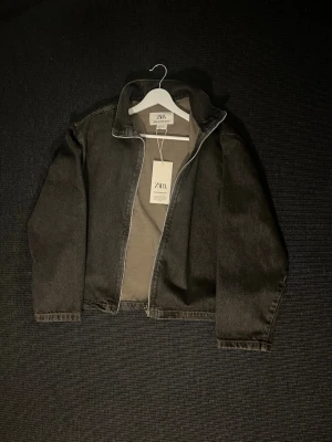 Svart jeansjacka från ZARA - Snygg svart jeansjacka från ZARA med klassisk krage och raka ärmar. Jackan har en stilren design utan synliga fickor framtill och stängs med dragkedja. Perfekt för dig som gillar en clean och modern look.