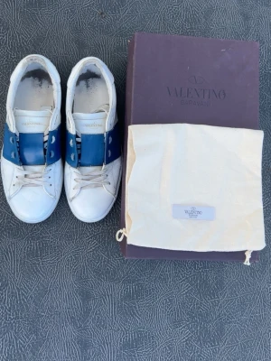 Valentino open blå - Snygga vita sneakers från Valentino med bred blå läderdetalj över snörningen. Klassisk låg modell med vita skosnören och diskret logga på plösen. Yttermaterial i läder och gummisula. Perfekta för dig som vill ha en stilren men ändå unik look. Storlek 42,5. Väldigt bra skick. Dustbag och box ingår. Enda dåliga är det ni kan se på bild 7. Men det är inget jag märker av och syns knappt. (Mina vänner har inte ens tänkt på det)  Pris kan diskuteras vid snabb affär! 🤝Självklart äkta. 