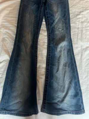 Blå utsvängda jeans med broderad ficka - Säljer ett par blå jeans med utsvängda ben och snygga detaljer. Jeansen har en broderad bakficka med kontrastsömmar i rött. De är lite slitna vid fötterna men annars väldigt fint skick.🩷 De är justerbara i midjan. 