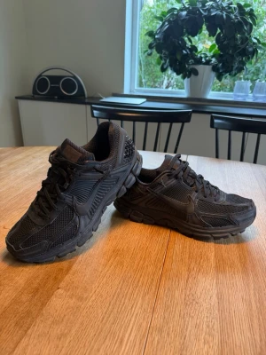 Helsvarta Nike Vomero skor.  - Säljer ett par helsvarta Nike Vomero skor med chunky sula och sportig design. Skorna har meshpaneler för bra ventilation och detaljer i syntetmaterial. Klassisk snörning och låg profil gör dem perfekta för en streetwear-look.
