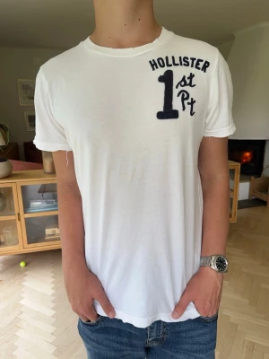 Hollister T-shirt vintage med tryck herr vit med slitningar - Vit t-shirt från Hollister i med coola slitningar och skön, mjuk kvalitet. Tryck med "Surf Flyers 1st Pt Hollister" på både bröstet och ryggen. Plagget har en snygg, medvetet lite sliten vintagelook i kanterna (raw edges) vid ärmar och krage. Perfekt till sommaren eller vardags! En fläck som visas på bild, annars gott skick.#hollister #tshirt #surf #y2k #vintage #california #streetwear #vittshirt #sommar