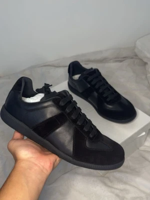 Maison margiela - Stilrena svarta sneakers med ovandel i både läder och mocka. Klassisk låg modell med svarta skosnören och diskreta detaljer. Perfekta för dig som gillar en clean och tidlös look. Snygg och bekväm sula som passar till många outfits.