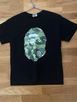 Svart t-shirt med camo-tryck - Säljer en svart t-shirt med stort kamouflagemönstrat tryck av ett ansikte både fram och bak. T-shirten har korta ärmar och en klassisk passform. Perfekt för dig som gillar streetwear och vill sticka ut med en unik design.