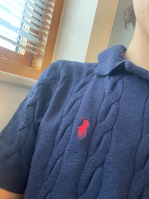 Marinblå kabelstickad pikétröja - Säljer en marinblå kabelstickad pikétröja med klassisk krage och korta ärmar. Tröjan har ett rött broderat logomärke på bröstet och är tillverkad i ett mjukt material som känns bekvämt mot huden. Perfekt för en stilren och avslappnad look.