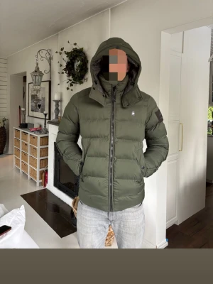 Olivgrön G-STAR Raw dunjacka med huva - Snygg äkta olivgrön G-STAR Raw dunjacka med huva och dragkedja framtill. Jackan har en quiltad design, hög krage och två sidofickor. Liten logotyp på bröstet och patch på ärmen. Perfekt för kalla vinterdagar och riktigt varm.