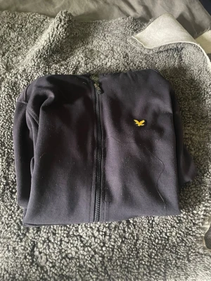 Lyle Scott hel zip tröja - Säljer en svart hoodie från Lyle & Scott med dragkedja framtill och den klassiska gula loggan på bröstet. Hoodien har huva med dragsko och två fickor framtill. Perfekt för en chill och stilren look.
