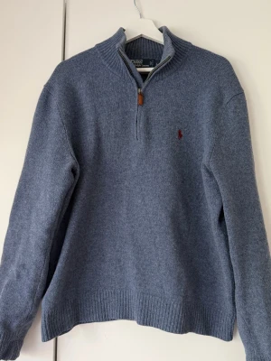 Blå halvzip tröja från Polo Ralph Lauren - Snygg blå stickad tröja från Polo Ralph Lauren med halvzip och ribbade muddar. Tröjan har en liten röd logga broderad på bröstet och är gjord i mjukt material. Perfekt till jeans eller chinos för en clean look.